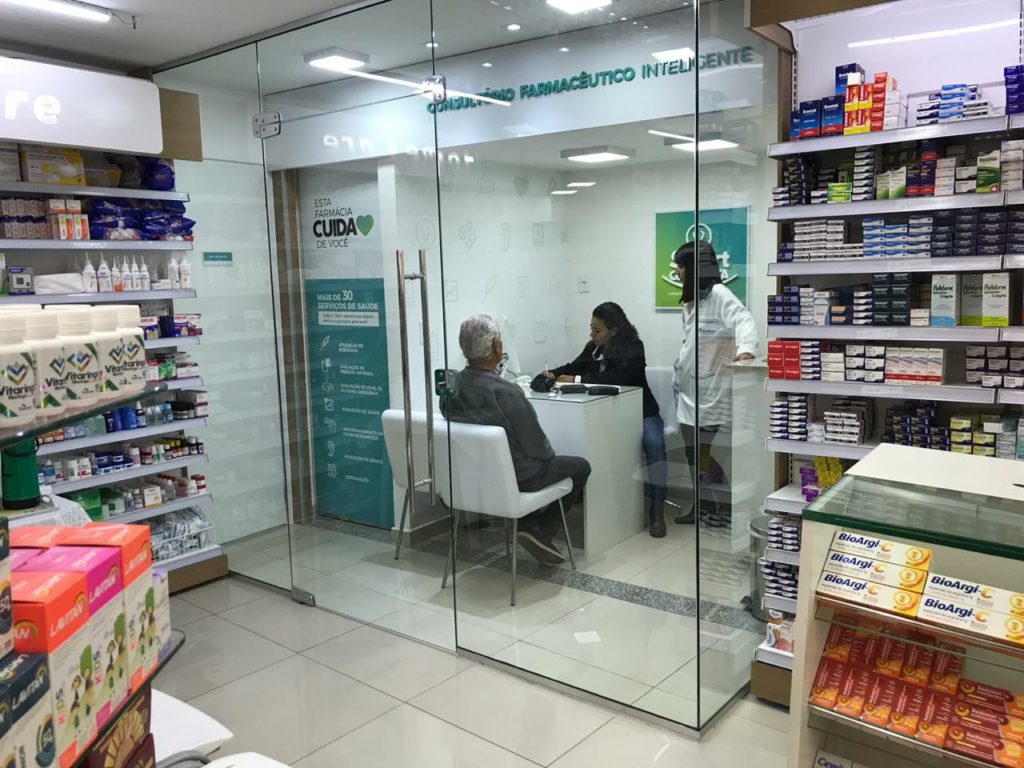 foto de um consultório farmacêutico dentro de uma farmácia, três pessoas dentro do consultório, um senhor e duas farmacêuticas