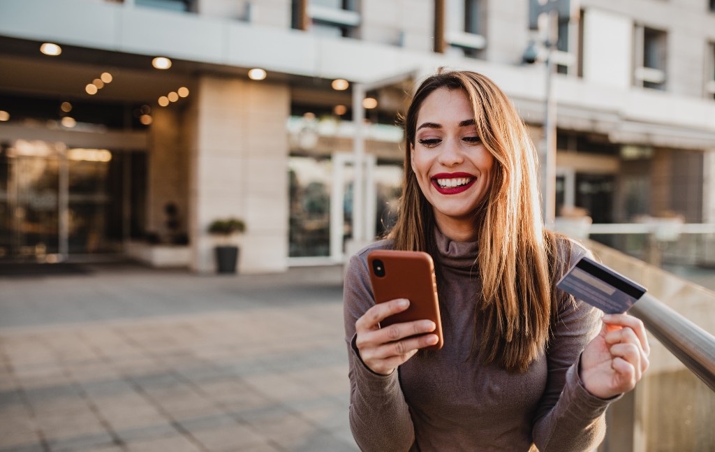mulher sorridente com celular e um cartão na mão efetuando uma compra