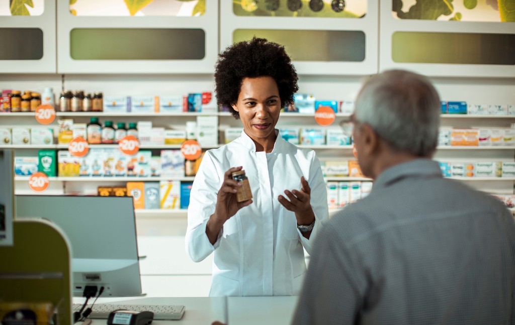 senhor de idade no balcão da farmácia recebendo intruções de farmacêutica que está segurando um frasco de remédio